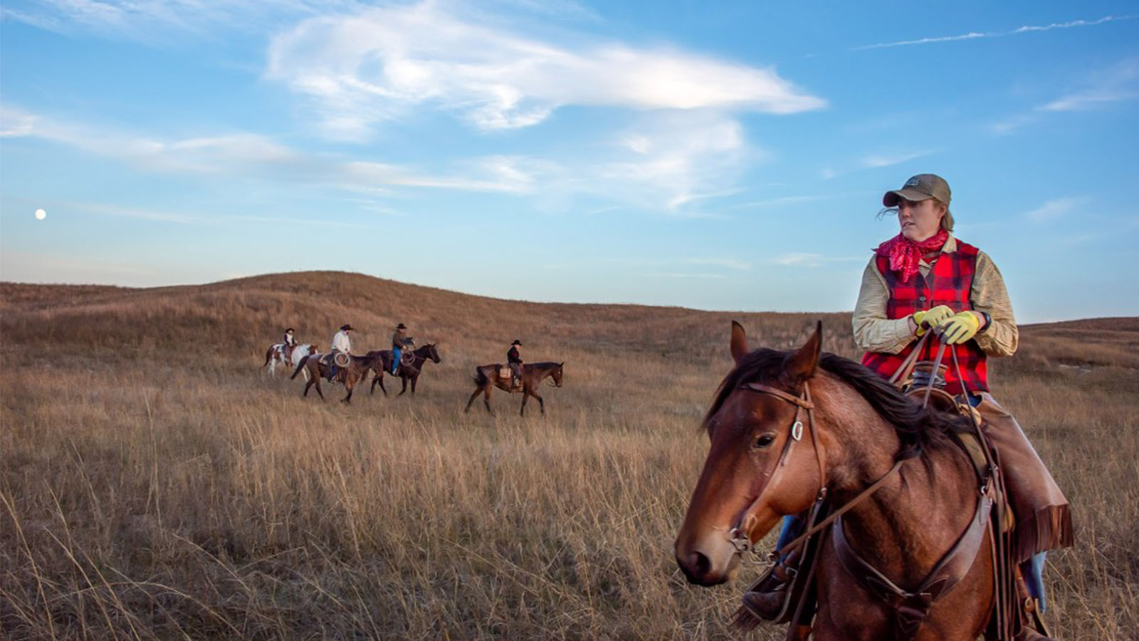 Riders on horseback traverse grassy plains under a blue sky with wispy clouds and a visible moon.