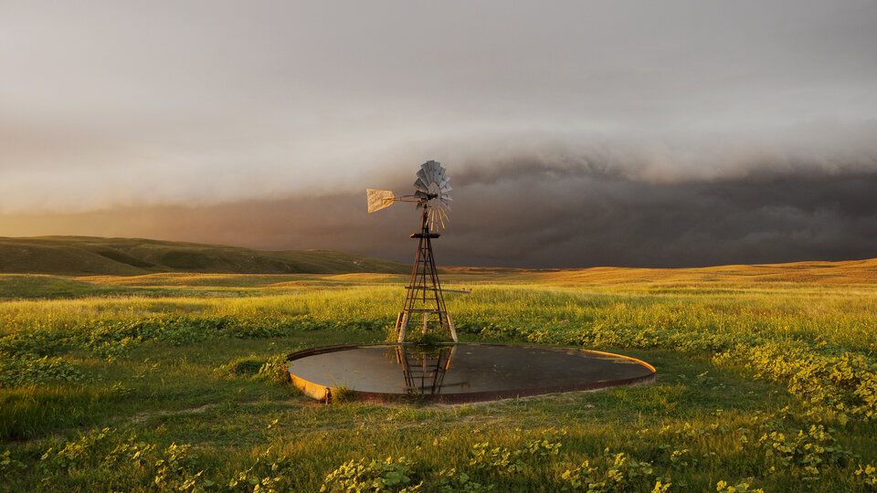 Windmill standing over a small pond in a grassy field under a cloudy, golden-lit sky.