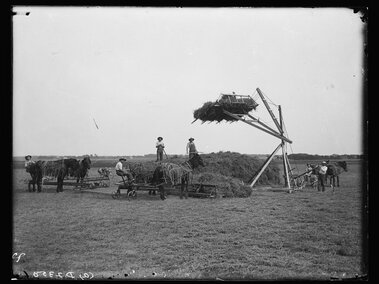 Farm workers loading hay onto a large stack using horse-drawn carts and a mechanical lift.