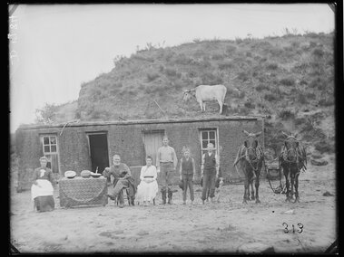 A family poses in front of a sod house with a cow on the roof and two horses beside them.