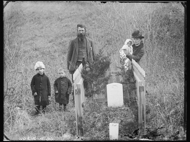 A family gathered at a fenced gravesite in a grassy field, one holding a young child.